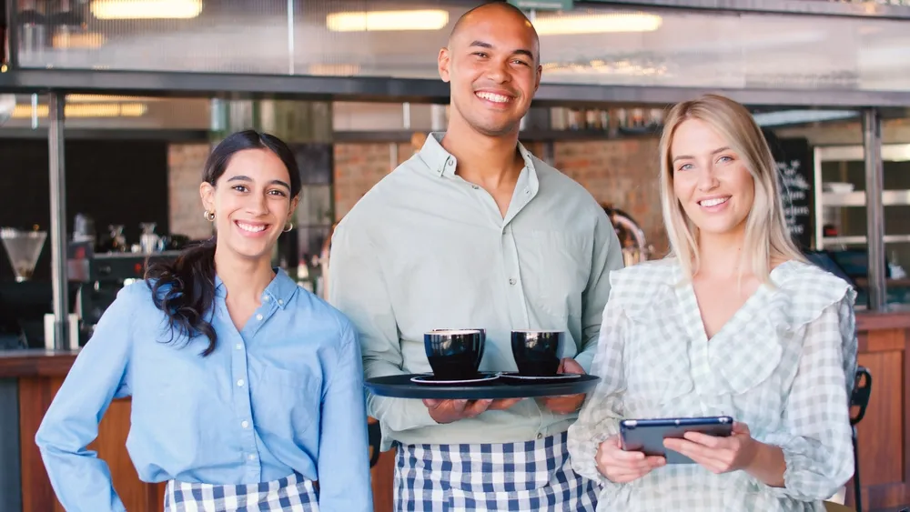 A smiling female restaurant owner, with members of her wait staff, standing in front of her restaurant with a tablet in hand, managing her online reviews on Google Reviews and Yelp.