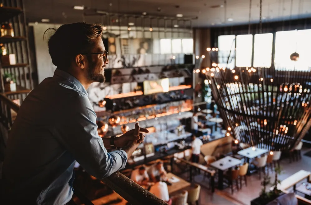 A restaurant owner leans against the railing of the second floor of his establishment as he looks across the main dining area. Digital marketing on TikTok for restaurants can help you reach new customers and grow your brand.