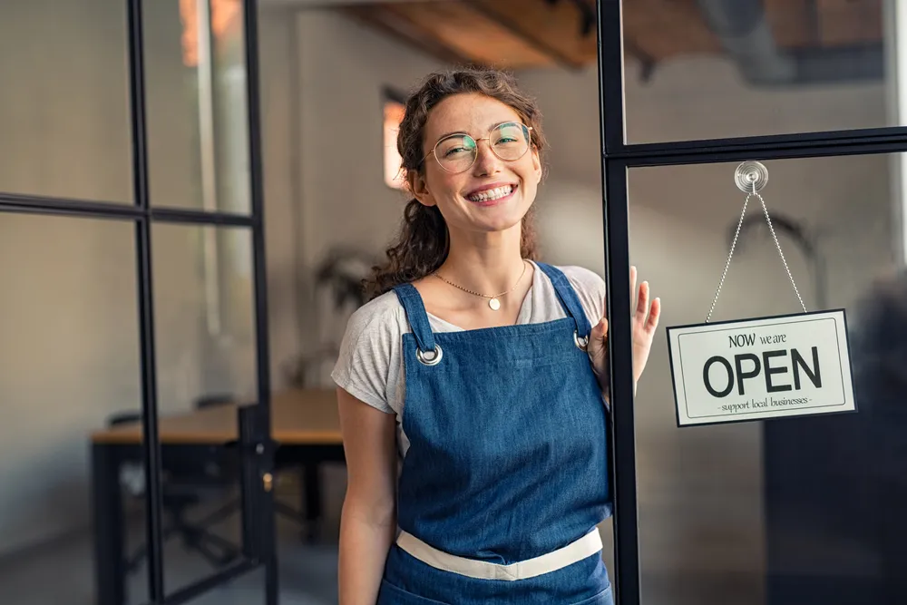 Una joven propietaria de un negocio de pie en la entrada de su restaurante con el cartel de 'Abierto Ahora' en la puerta. Su éxito es un testimonio del poder de las estrategias de marketing digital efectivas.