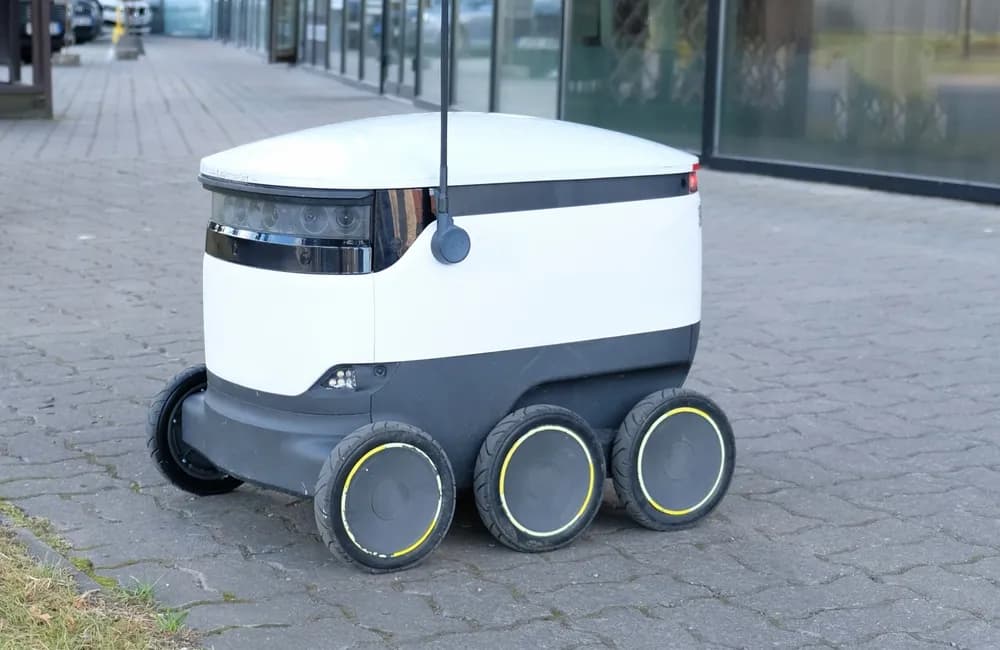 Profile view of a food delivery robot on a city sidewalk, with buildings and trees in the background. The box-shaped operators are designed to navigate sidewalks and streets, delivering food directly to customers.