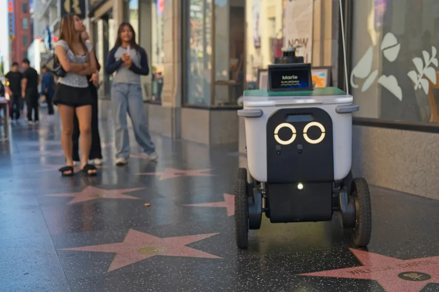 A food delivery robot rolls down landmark sidewalk of Hollywood Boulevard, with the iconic Hollywood stars embedded in the sidewalk. Despite being a highly trafficked area, the robot is able to navigate the area autonomously.