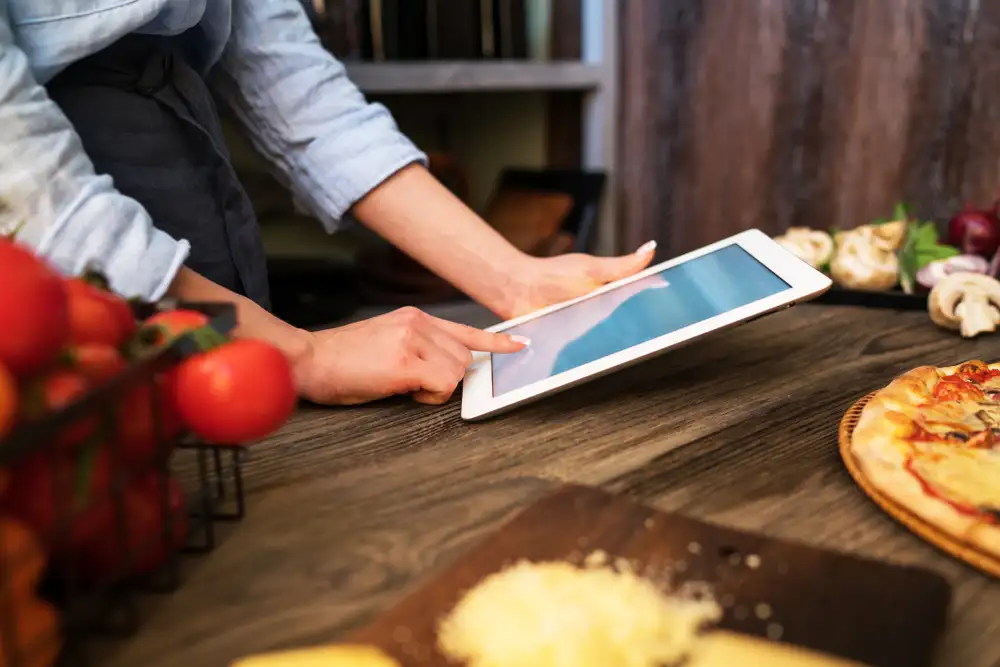 Se puede usar una calculadora de costos de alimentos en línea en tabletas, computadoras portátiles o de escritorio, lo que facilita la administración de los costos de alimentos en cualquier entorno de cocina, como se muestra en esta imagen de un gerente de restaurante que usa una tableta para monitorear cómo calcular el costo de los alimentos.