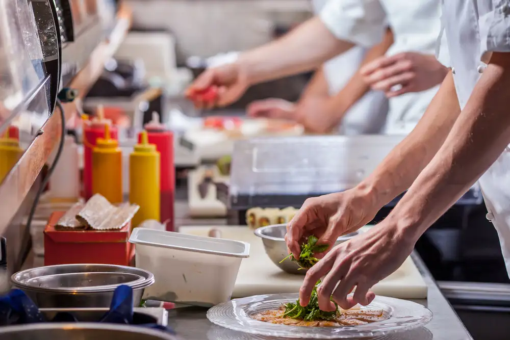 Los restaurantes deben controlar constantemente los costos de los alimentos para garantizar su rentabilidad. Esta imagen muestra a cocineros preparando comida en la cocina de un restaurante con diversos ingredientes, lo que resalta la importancia de un cálculo eficaz de los costos de los alimentos.
