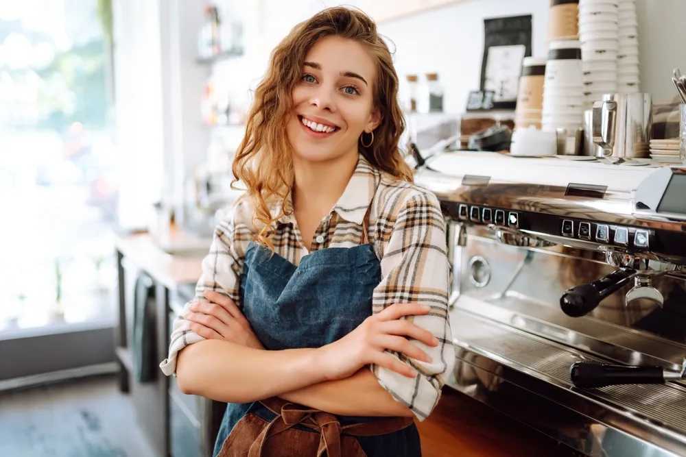 A cafe owner leaning against her counter smiling, with a coffee machine and pastries in the background. Follow our practical tips to start your own cafe business successfully.