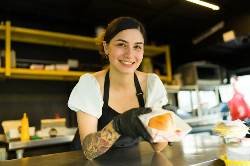 A smiling food truck owner serves a sandwich. No need to keep dreaming, start your food truck business in Providence RI today!