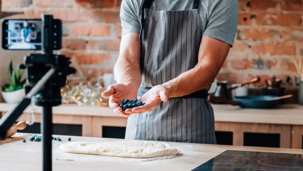 A culinary instructor in front of a camera teaching an online cooking class, with a laptop and professional kitchen setup.