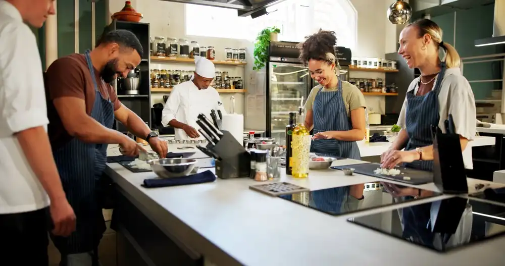 Culinary students in a live cooking school class, following along with an instructor demonstrating techniques in a professional kitchen.