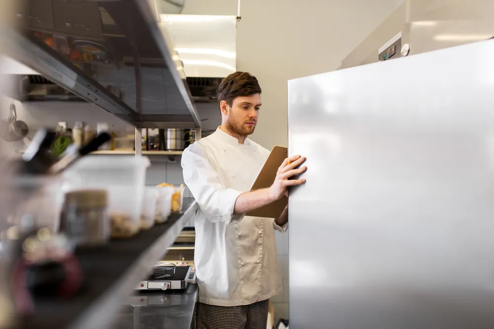 A kitchen manager checking the temperature of a commercial refrigerator. Buying used commercial kitchen equipment like refrigerators can be risky due to potential maintenance issues.