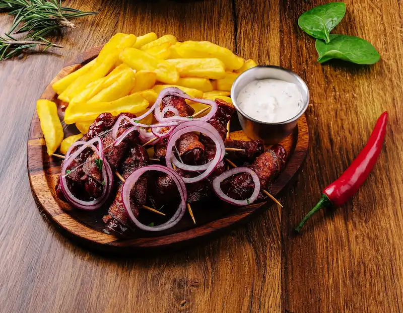Un plato de carne a la parrilla en brocheta, papas fritas y cebolla morada. Los colores de cada elemento del plato resaltan, dándole un aspecto apetitoso y atractivo. Las imágenes del menú deben aprovechar los colores del plato para hacerlo más atractivo.