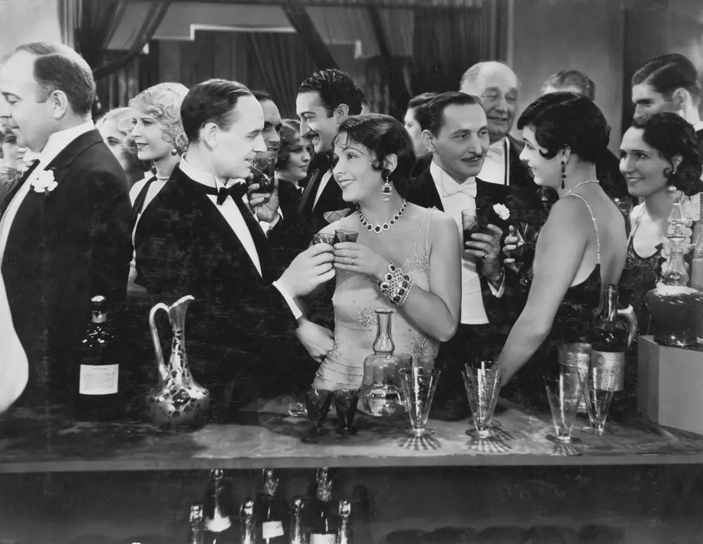 Bartenders and mixologists are often confused, but they are not the same. A mixologist is a bartender who specializes in creating unique cocktails and drinks. In this vintage black and white photo, guests are enjoying cocktails at a bar.