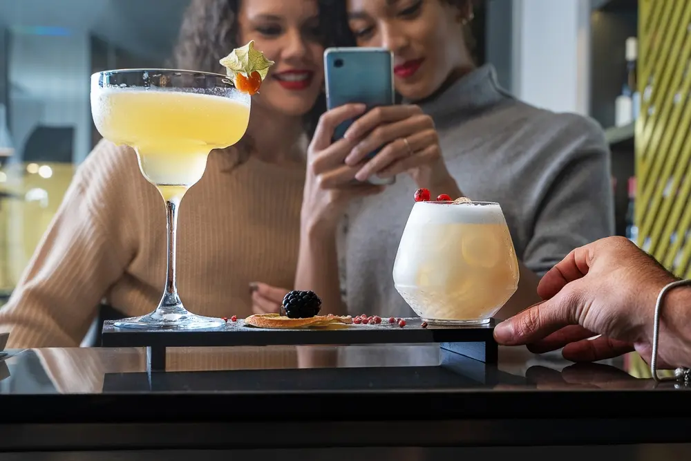 Understanding what is a mixologist is important for bars and restaurants. A trained mixologist knows how to keep the quality of drinks the same every time, ensuring customers get great drinks as shown in this image of two female friends taking a photo of their drinks for posting to Instagram.